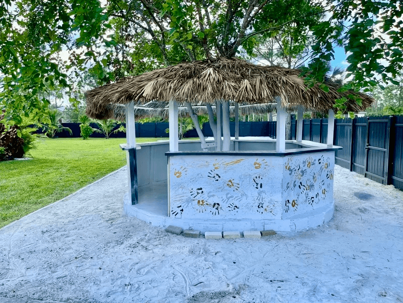 Outdoor hut with thatched roof, handprints on white walls, surrounded by greenery and sand.